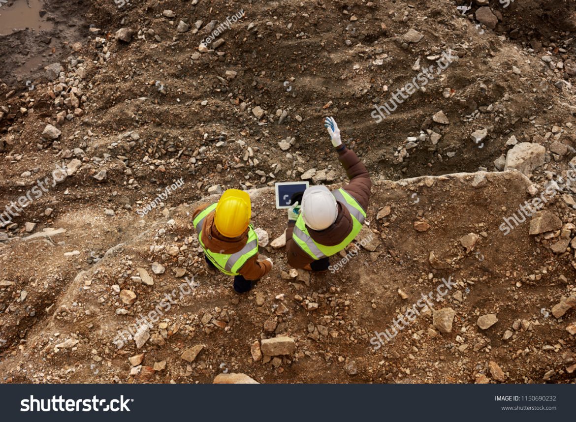 stock-photo-above-view-shot-of-two-industrial-workers-wearing-reflective-jackets-standing-on-mining-worksite-1150690232